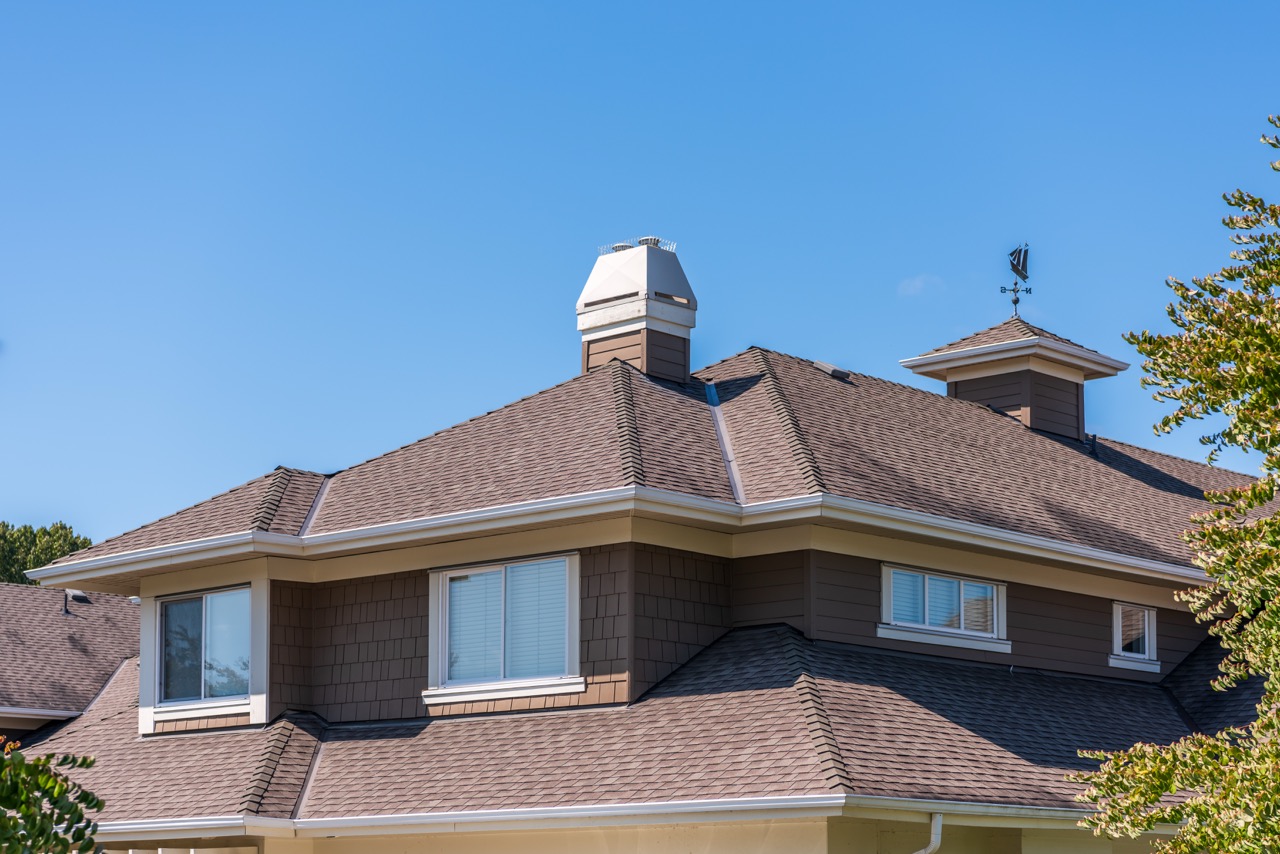 Completed residential hip roof with brown asphalt shingles, white-capped chimney and a cupola.