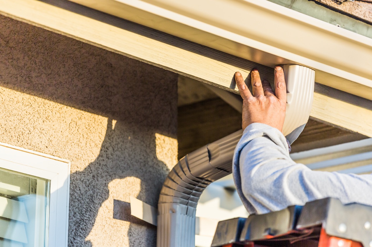 Worker's hand holding a downspout elbow against the fascia while installing rain gutters on a stucco home.