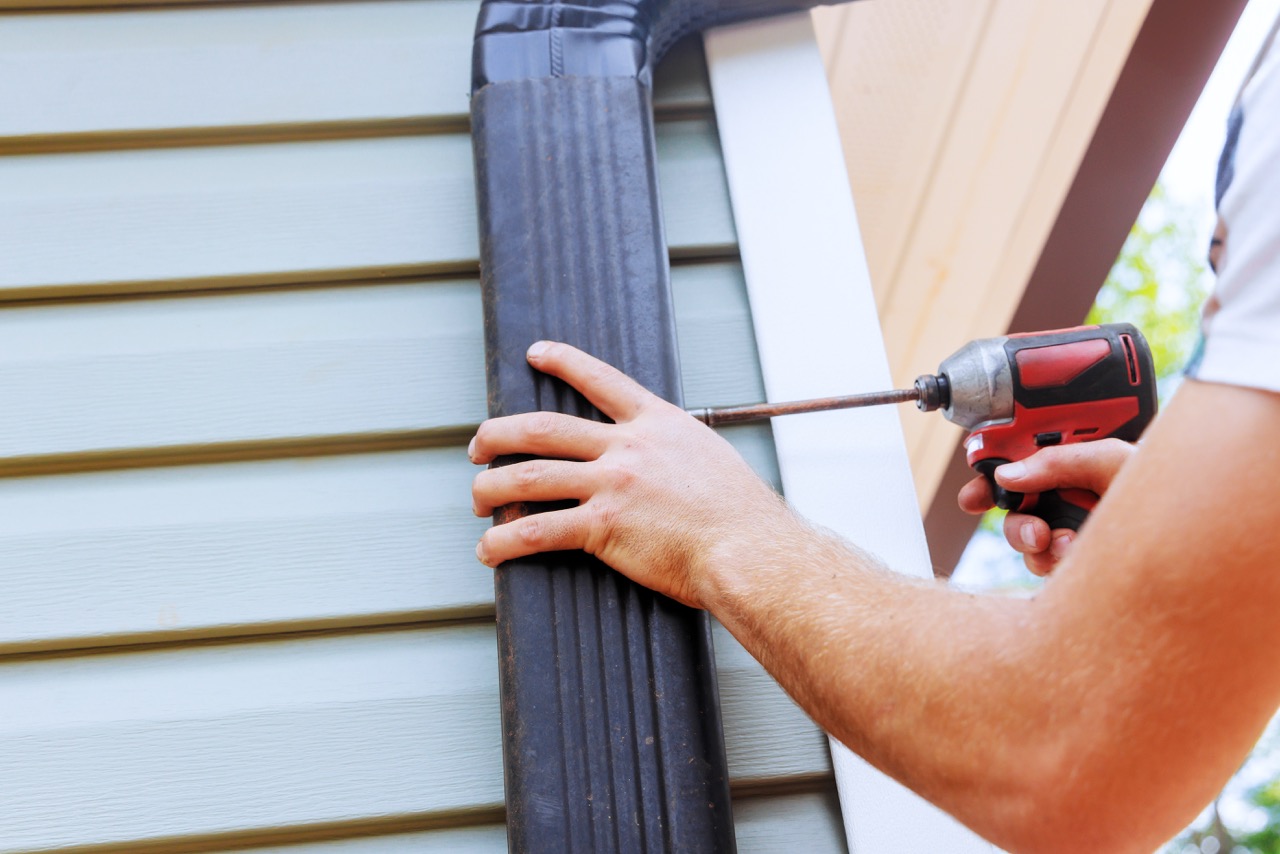 Worker fastening a black downspout to light green vinyl siding with a cordless impact driver.