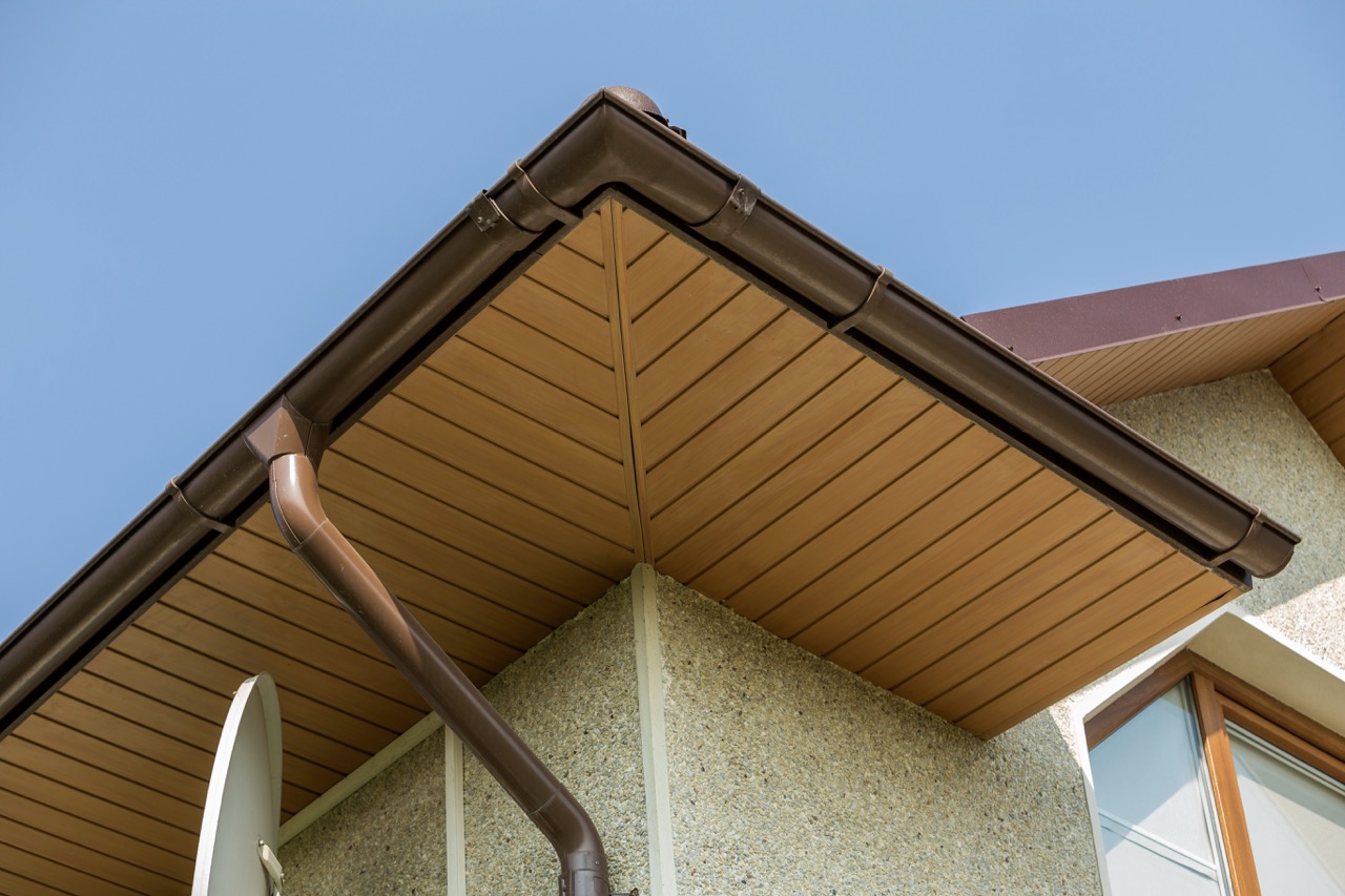 Roof corner on a New Jersey home showing wood-look soffit panels, brown gutters and a downspout against a clear sky.