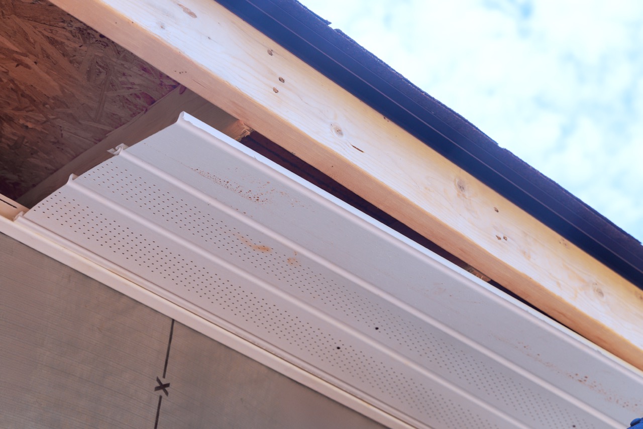 Partially installed white perforated vinyl soffit panel along a roof eave with exposed fascia board and roof sheathing.